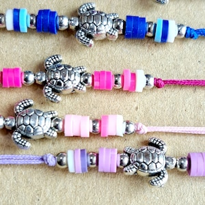 Puede incluir: Cuatro pulseras coloridas con dijes de tortuga plateada. Cada pulsera presenta un esquema de color diferente con pequeñas cuentas redondas. Las pulseras están hechas con una variedad de colores, incluyendo rosa, azul, morado y blanco.