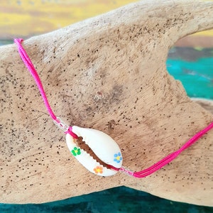 Peut inclure: Un bracelet en cordon rose avec un charme en coquillage blanc. Le coquillage est peint avec des fleurs colorées.