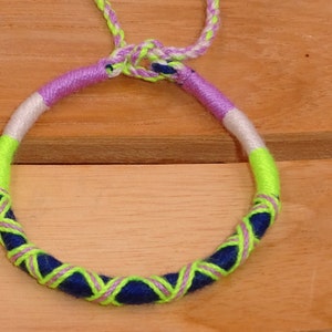 Peut inclure: Un bracelet d'amitié coloré fait de fils tissés de couleur violette, blanche, verte et bleue.
