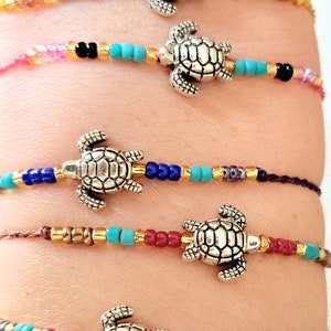 Puede incluir: Cinco pulseras de cuentas de colores con dijes de tortuga plateada. Cada pulsera presenta una combinación de colores de cuentas diferente, incluyendo turquesa, rosa, rojo, azul y dorado.