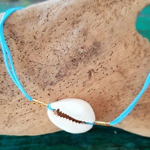 May include: A delicate necklace featuring a white seashell pendant. The shell is strung on a vibrant, light blue cord, accented with gold-colored metal beads. The necklace is displayed on a piece of driftwood, creating a beachy aesthetic.