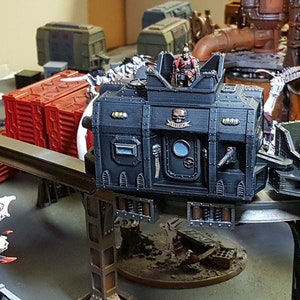 May include: A black and grey miniature model of a futuristic vehicle with a skull and crossbones symbol on the side. The vehicle is on a track and has a figure in the driver's seat.