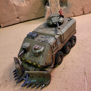 Puede incluir: Un vehículo militar verde y marrón con una gran cuchilla de metal en la parte delantera. El vehículo tiene una torreta en la parte superior con un cañón apuntando hacia adelante. El vehículo está sentado sobre una superficie arenosa.