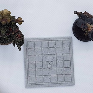 May include: A grey square tile with a grid pattern and a skull design in the centre. The tile is surrounded by a white background.
