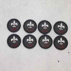 Peut inclure: Huit patchs thermocollants ronds noirs avec un motif de fleur de lys blanche et le texte "1CP" en rouge.