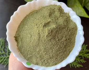 May include: A white, scalloped bowl filled with finely ground, green powder. The bowl is held in a hand, with green sprigs and a dark surface in the background. The powder appears to be a culinary herb or spice.