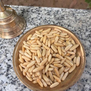 Puede incluir: Un pequeño plato redondo de latón antiguo lleno de piñones. Las nueces son de color beige claro y de forma ovalada. El plato se encuentra sobre una superficie moteada gris y blanca, con un objeto de latón en el fondo.