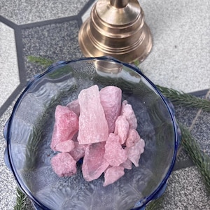 May include: A clear glass bowl with a blue rim filled with pink crystals. A gold-colored bell is in the background. The bowl sits on a gray and white tiled surface, with a few sprigs of greenery.