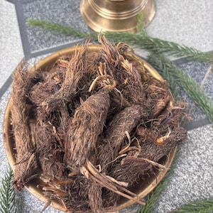May include: A brass bowl filled with dried, brown roots. A small brass bell sits in the background, with green sprigs of foliage around the bowl. The roots have a fibrous texture.