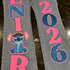May include: Light blue denim jeans featuring glittery pink and blue lettering that spells out SENIOR 2026. The design includes two graphic illustrations of the character Stitch wearing a black graduation cap and holding a diploma.