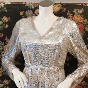 Könnte beinhalten: Ein langärmeliges, silbernes Paillettenkleid mit V-Ausschnitt und geometrischem Muster. Das Kleid hat einen Bindegürtel in der Taille.