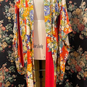 Könnte beinhalten: Ein farbenfrohes Blumenkimono mit rotem Futter wird auf einer Schneiderpuppe präsentiert. Das Kimono hat ein lebendiges Muster aus gelben, orangefarbenen, blauen und weißen Blumen. Auf der Schneiderpuppe steht der Text "Fair Lady".