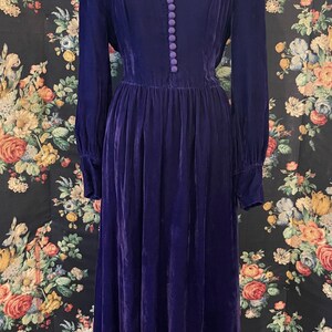 Puede incluir: Un vestido largo de terciopelo morado oscuro con mangas largas y una fila de botones en la parte delantera. El vestido tiene cintura alta y falda amplia. El escote est&aacute; ribeteado con una tela morada m&aacute;s clara. El vestido se exhibe en un maniqu&iacute;.