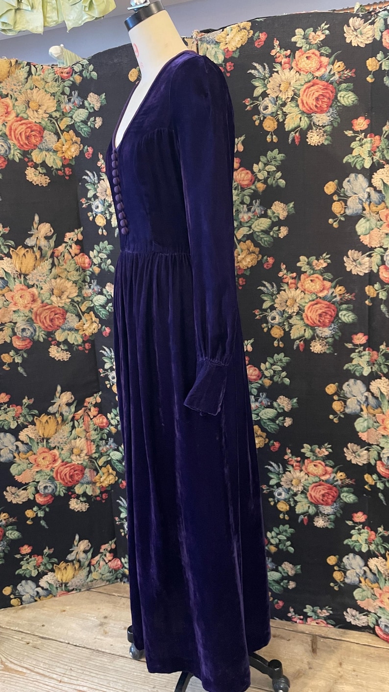 Puede incluir: Un vestido largo de terciopelo morado oscuro con mangas largas y escote en V. El vestido presenta una serie de peque&ntilde;os botones en la parte delantera y una falda fruncida. El vestido se exhibe en un maniqu&iacute; contra un fondo floral.