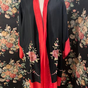 Könnte beinhalten: Schwarzer Kimono-Stil Robe mit roter Verzierung und floraler Stickerei. Die Robe hat ein fließendes Design mit weiten Ärmeln und einem kontrastierenden roten Rand. Die Stickerei umfasst rosa Rosen und grüne Blätter. Die Robe ist etwa 150 cm lang.