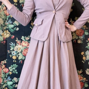 Könnte beinhalten: Ein Vintage-Anzug in Lavendel. Die Jacke hat einen einzelnen Knopfverschluss und zwei Vordertaschen. Der Rock hat eine passende Lavendelfarbe und ein plissiertes Design. Der Anzug ist aus einem weichen Stoff gefertigt und eignet sich für formelle Anlässe.