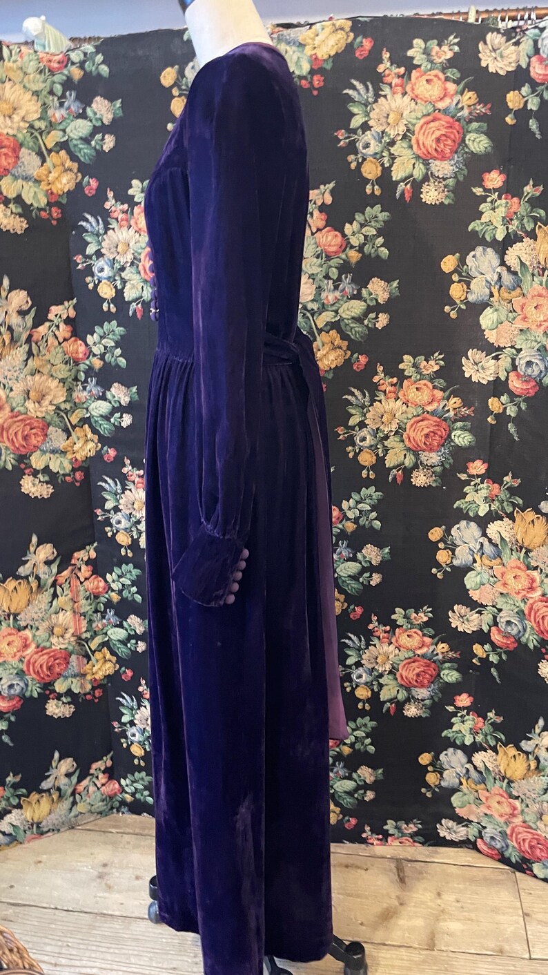 Puede incluir: Un vestido largo de terciopelo morado oscuro con mangas largas y un corpi&ntilde;o ajustado. El vestido presenta detalles de botones en los pu&ntilde;os y un cintur&oacute;n a juego. El vestido se exhibe en un maniqu&iacute; sobre un fondo floral.