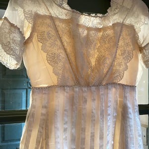 Puede incluir: Un vestido vintage blanco con un corpiño de encaje y una falda a rayas. El vestido tiene un escote redondo y mangas cortas.