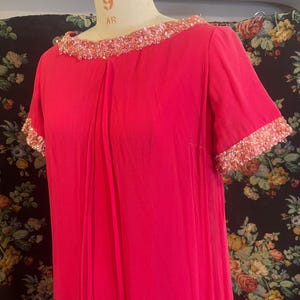 May include: A vibrant fuchsia-colored dress with short sleeves and a round neckline. The neckline and sleeve edges are embellished with a band of shimmering, light-colored sequins. The dress is displayed on a mannequin against a floral patterned backdrop.