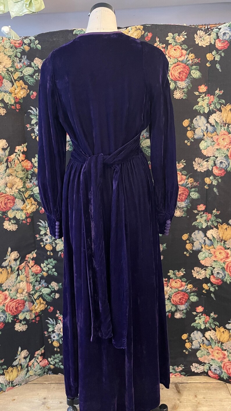 Puede incluir: Un vestido largo de terciopelo morado oscuro con mangas largas y un cintur&oacute;n a juego atado a la cintura. El vestido tiene cuello en V y pu&ntilde;os abotonados. El vestido se exhibe en un maniqu&iacute; sobre un fondo floral.