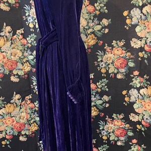 Puede incluir: Un vestido largo de terciopelo morado oscuro, con mangas largas y cintur&oacute;n a juego. El vestido presenta un cuello alto y botones decorativos en los pu&ntilde;os. El vestido se exhibe en un maniqu&iacute; sobre un fondo floral.