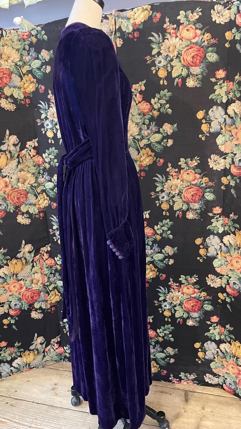 Puede incluir: Un vestido largo de terciopelo morado oscuro, con mangas largas y cintur&oacute;n a juego. El vestido presenta un cuello alto y botones decorativos en los pu&ntilde;os. El vestido se exhibe en un maniqu&iacute; sobre un fondo floral.