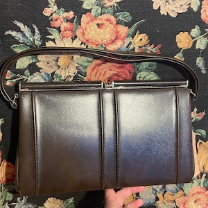 Könnte beinhalten: Eine braune Leder-Clutch mit einem silbernen Verschluss und einem einzigen Riemen.