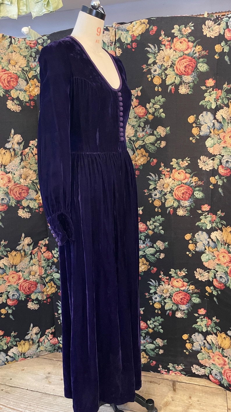 Puede incluir: Un vestido largo de terciopelo morado oscuro con mangas largas y escote en V. El vestido presenta una fila de botones en la parte delantera y se exhibe en un maniqu&iacute;. El fondo es un estampado floral.