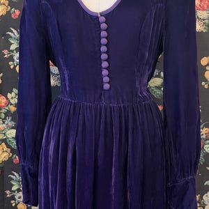 Puede incluir: Vestido largo de terciopelo morado oscuro con escote en V ribeteado en un morado m&aacute;s claro. El vestido presenta una serie de botones a juego en la parte delantera y una falda fruncida. Las mangas tienen un ligero abullonado en el hombro y pu&ntilde;os ajustados.