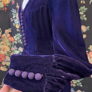 Puede incluir: Un vestido de terciopelo morado oscuro con escote en V y mangas largas. El vestido presenta una fila de peque&ntilde;os botones a juego en la parte delantera y botones de bola decorativos en los pu&ntilde;os. La tela tiene un aspecto suave y lujoso.