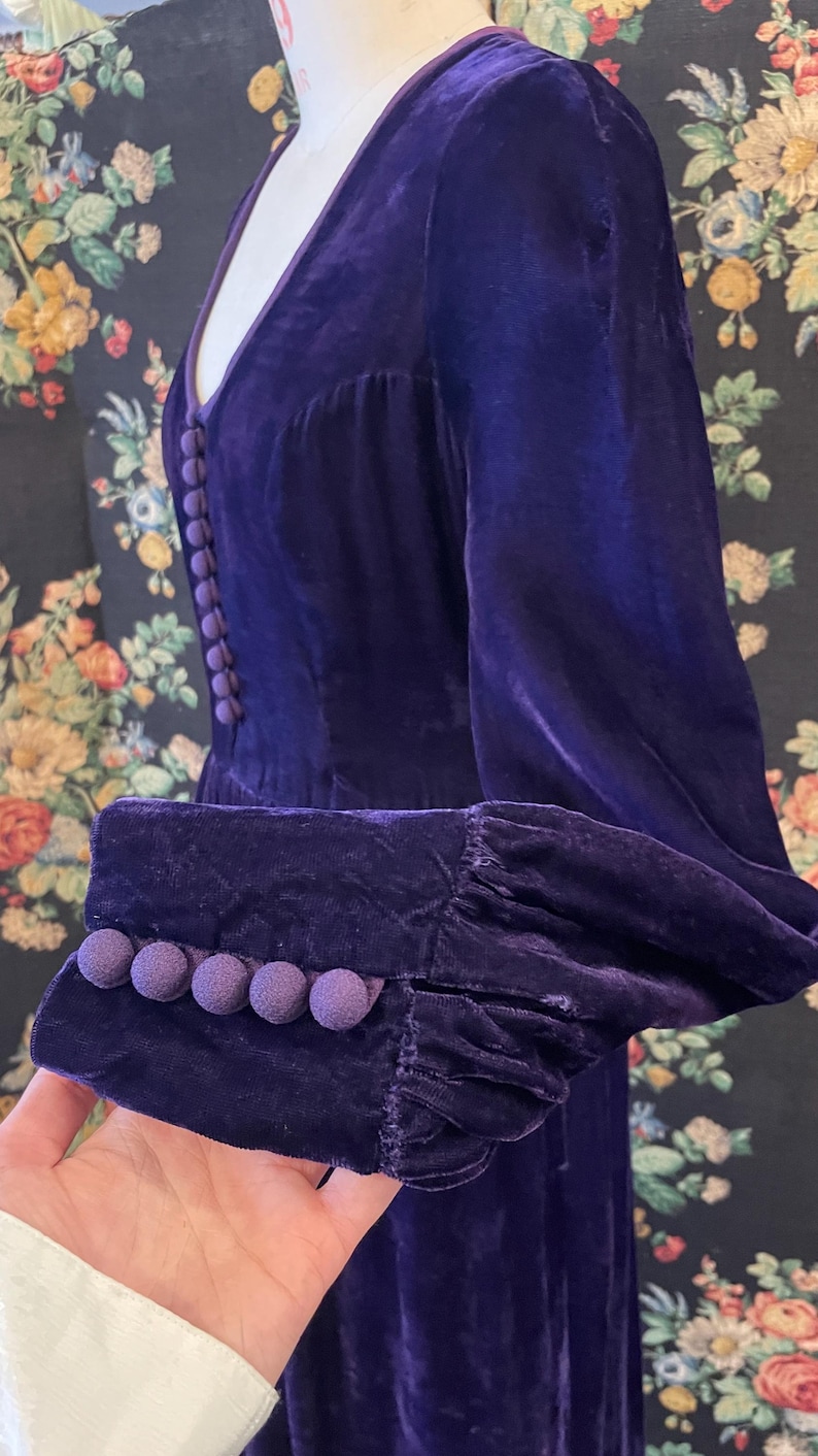 Puede incluir: Un vestido de terciopelo morado oscuro con escote en V y mangas largas. El vestido presenta una fila de peque&ntilde;os botones a juego en la parte delantera y botones de bola decorativos en los pu&ntilde;os. La tela tiene un aspecto suave y lujoso.