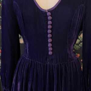 Puede incluir: Un vestido vintage de terciopelo morado oscuro de manga larga. El vestido presenta un escote en V ribeteado con una tela morada m&aacute;s clara y una fila de botones a juego en la parte delantera. La falda es plisada y el vestido se exhibe en un maniqu&iacute;.