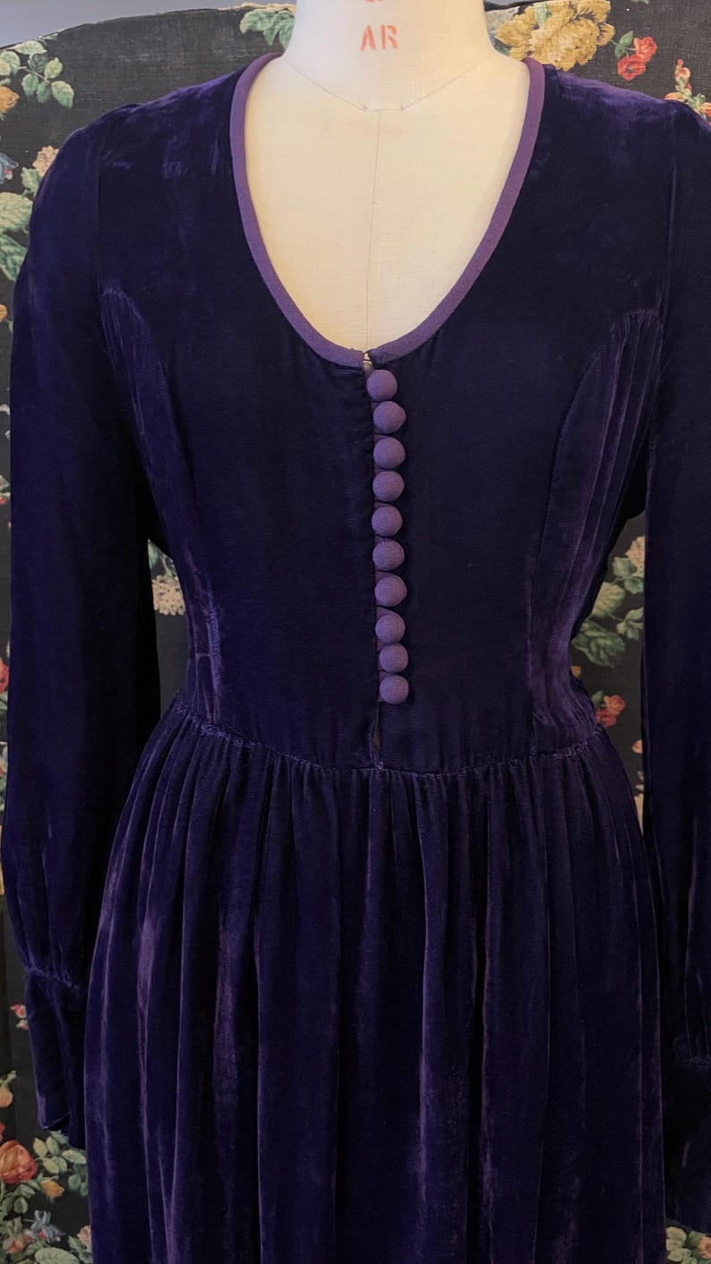 Puede incluir: Un vestido vintage de terciopelo morado oscuro de manga larga. El vestido presenta un escote en V ribeteado con una tela morada m&aacute;s clara y una fila de botones a juego en la parte delantera. La falda es plisada y el vestido se exhibe en un maniqu&iacute;.