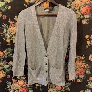 Könnte beinhalten: Ein silberner, glitzernder Cardigan mit V-Ausschnitt, langen Ärmeln und zwei Taschen. Der Cardigan hat einen Knopfverschluss und ein Etikett mit der Aufschrift "Ross Stop".