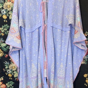 May include: A light purple open-front cardigan with a floral pattern and pink trim. The cardigan has long sleeves and a tie closure. The fabric has a subtle dotted design and a decorative pattern along the bottom.