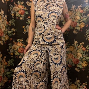 Può includere: Un completo a due pezzi composto da un top senza maniche e pantaloni a gamba larga. L'ensemble presenta un motivo floreale ripetuto in beige, blu navy e pesca. Il top ha uno scollo alto. I pantaloni hanno un design a gamba larga.