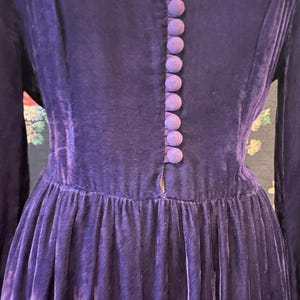 Puede incluir: Un vestido vintage de manga larga en un tono morado intenso. El vestido presenta un escote en V ribeteado en un morado m&aacute;s claro, una fila de botones cubiertos de tela a juego y una falda plisada. La tela parece ser terciopelo.