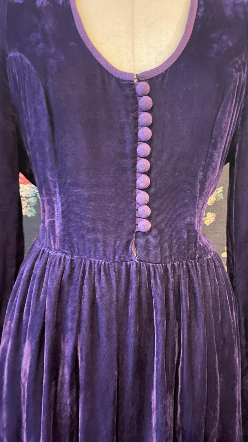 Puede incluir: Un vestido vintage de manga larga en un tono morado intenso. El vestido presenta un escote en V ribeteado en un morado m&aacute;s claro, una fila de botones cubiertos de tela a juego y una falda plisada. La tela parece ser terciopelo.