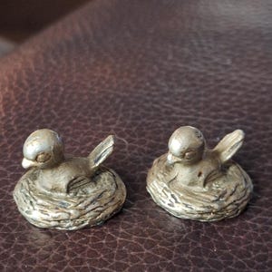 Puede incluir: Dos pequeñas figuras de pájaros de latón de estilo antiguo. Cada pájaro está posado sobre una base en forma de nido, con las alas ligeramente extendidas. Los pájaros tienen un aspecto detallado y texturizado, sobre un fondo marrón.