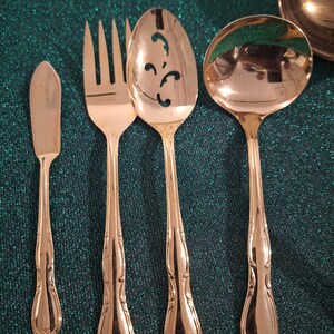 Puede incluir: Un juego de cubiertos dorados, que incluye un cuchillo de mantequilla, un tenedor, una cuchara ranurada y un cucharón. La cubertería tiene detalles ornamentales en los mangos y está dispuesta sobre una superficie de color turquesa. La cuchara ranurada tiene un diseño decorativo.