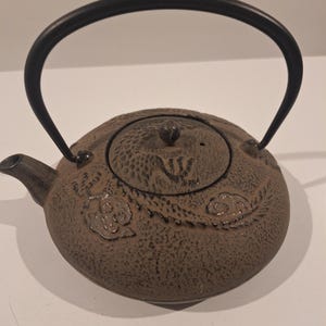 Op de afbeelding: Een bruine gietijzeren theepot met een zwarte handgreep en tuit. De theepot heeft een textuur en een deksel met een kleine knop. Decoratieve bloemen- en wolkenpatronen zijn op het oppervlak van de theepot geëmbosseerd.