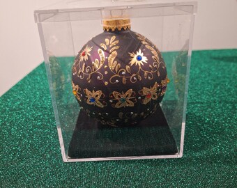 May include: A dark purple Christmas ornament with gold floral designs and colorful jewel accents. The ornament is displayed in a clear plastic box on a black base, set against a green, glittery surface. The ornament's cap is gold.