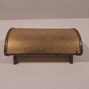 Puede incluir: Un pequeño cofre de latón antiguo con la parte superior curvada y adornos de latón. El cofre es rectangular y descansa sobre cuatro patas cortas y rectangulares. El latón tiene un aspecto ligeramente envejecido con algunos arañazos en la superficie.