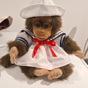 May include: A plush monkey doll with brown fur and tan hands and feet, wearing a white sailor-style outfit with a matching white hat. The outfit features a navy blue striped collar and a bright red ribbon tied in a bow at the center of the chest.