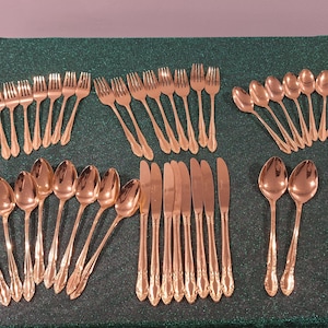 Puede incluir: Una colección de cubiertos dorados, que incluye tenedores, cucharas y cuchillos, dispuestos sobre una superficie verde. La cubertería tiene un diseño decorativo en los mangos. La disposición sugiere un entorno de comedor formal.