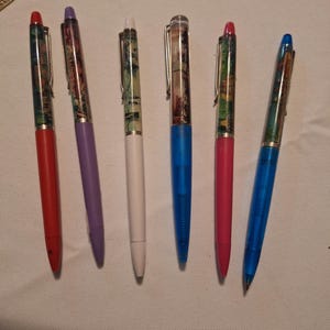 Puede incluir: Seis bolígrafos de colores. Los bolígrafos tienen un diseño de paisaje en el cuerpo y son rojos, morados, blancos, azules y rosas. Cada bolígrafo tiene una punta del mismo color y un clip plateado.