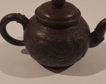 May include: A small, brown teapot with a rounded body, a curved handle, and a spout. The teapot features a detailed dragon carving on its side and a small, round knob on the lid. The teapot is made of a dark brown material.