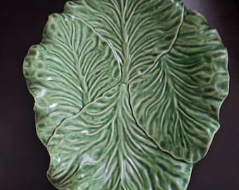 Puede incluir: Una fuente de cerámica verde con forma de hoja grande. La fuente tiene vetas detalladas y un acabado brillante. El diseño en forma de hoja es una pieza única y decorativa para servir alimentos o como objeto decorativo.