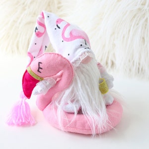 May include: A pink flamingo gnome with a white beard and a pink and white flamingo patterned hat. The gnome is sitting on a pink flamingo float.
