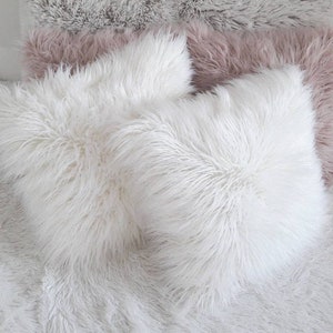 May include: Three fluffy throw pillows, one white and two in a light pink color, are arranged on a white fluffy surface. The pillows are made of a soft, plush material.